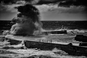 grayscale photo of man standing on dock near sea waves
