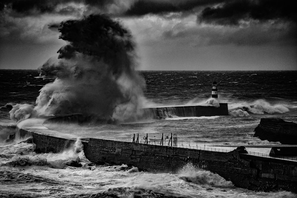 grayscale photo of man standing on dock near sea waves