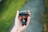 A hand is holding a small figurine wearing a cap, sunglasses, and black clothing. The figurine appears to have stylized features typical of collectible figures. The background is a blurred outdoor setting with green grass and a gray pathway.