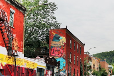 A colorful urban street scene features buildings adorned with vibrant graffiti art. One prominent red brick building displays a large mural of a fantastical creature. The setting includes several other buildings with graffiti, trees providing greenery, and a clear sky. Fire escapes are visible on the building walls.