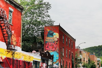 A colorful urban street scene features buildings adorned with vibrant graffiti art. One prominent red brick building displays a large mural of a fantastical creature. The setting includes several other buildings with graffiti, trees providing greenery, and a clear sky. Fire escapes are visible on the building walls.