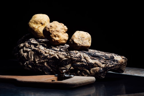 Three large truffles are artfully arranged on a piece of textured wood placed on a surface. The background is dark, highlighting the earthy tones of the truffles and the wood.