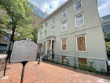 A historical building with a sign titled 'White House of the Confederacy' in front. The building is three stories tall with boarded-up windows on the ground floor. It has a traditional architectural style with white trim and light green paint. The surrounding area includes a red brick sidewalk, wrought iron fence, and lush green trees. Modern buildings are visible in the background.