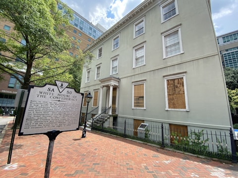 A historical building with a sign titled 'White House of the Confederacy' in front. The building is three stories tall with boarded-up windows on the ground floor. It has a traditional architectural style with white trim and light green paint. The surrounding area includes a red brick sidewalk, wrought iron fence, and lush green trees. Modern buildings are visible in the background.