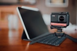 A digital camera mounted on a tripod sits on a wooden table in front of a tablet positioned on a keyboard case. The background is softly blurred, creating a focus on the camera and tablet.