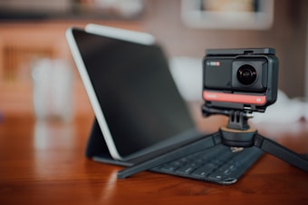 A digital camera mounted on a tripod sits on a wooden table in front of a tablet positioned on a keyboard case. The background is softly blurred, creating a focus on the camera and tablet.