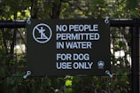 A sign is displayed on a metal fence with foliage in the background. The sign has a dark green background with white text that reads 'No People Permitted In Water For Dog Use Only.' There is also a graphic of a person walking on an incline with a prohibition symbol over it, and a small NYC Parks logo in the lower right corner.