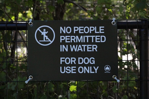 A sign is displayed on a metal fence with foliage in the background. The sign has a dark green background with white text that reads 'No People Permitted In Water For Dog Use Only.' There is also a graphic of a person walking on an incline with a prohibition symbol over it, and a small NYC Parks logo in the lower right corner.