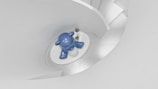 A polished blue sculpture resembling an animal is placed on a white platform at the bottom of a spiral staircase. The staircase is part of a minimalist, modern architectural design featuring smooth, flowing curves and light grey tones. A small decorative plant is placed beside the sculpture.