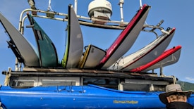 A collection of colorful kayaks and surfboards are mounted on a rack atop a vehicle. The kayaks come in shades of blue, green, and red, and the surfboards are positioned above them. The scene includes a clear blue sky and a sunlit atmosphere, contributing to a vibrant outdoor setting. A person wearing a wide-brimmed hat is visible at the lower right corner, gazing at the equipment.