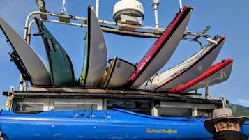 A collection of colorful kayaks and surfboards are mounted on a rack atop a vehicle. The kayaks come in shades of blue, green, and red, and the surfboards are positioned above them. The scene includes a clear blue sky and a sunlit atmosphere, contributing to a vibrant outdoor setting. A person wearing a wide-brimmed hat is visible at the lower right corner, gazing at the equipment.