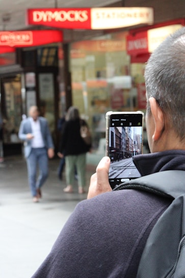 A person is photographing a street scene with a smartphone while standing in a busy shopping area. Blurred figures of pedestrians move in the background, and above them, a red-lit stationery shop sign is visible.