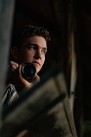 A person holding a camera appears partially illuminated, looking thoughtfully into the distance. The setting seems dimly lit and suggests a contemplative or focused atmosphere.