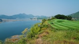 A scenic landscape shows a well-maintained golf course bordered by lush, green grass and a large lake. The background features mountains under a clear, blue sky. A piece of green maintenance equipment is visible in the foreground.