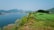 A scenic landscape shows a well-maintained golf course bordered by lush, green grass and a large lake. The background features mountains under a clear, blue sky. A piece of green maintenance equipment is visible in the foreground.