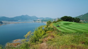 A scenic landscape shows a well-maintained golf course bordered by lush, green grass and a large lake. The background features mountains under a clear, blue sky. A piece of green maintenance equipment is visible in the foreground.