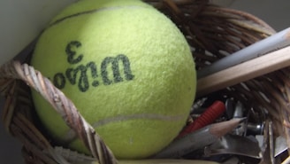 A tennis ball with the number 3 and brand name printed on it is placed inside a woven basket. Surrounding the ball are various stationery items including pencils, a ruler, and metal clips.