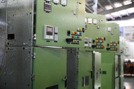 A row of industrial control panels painted in green with various dials, switches, and meters. The panels are aligned under a metal roof, and there are cables visible on one side. Brightly colored buttons in red, blue, yellow, and green are prominently placed on the panels.