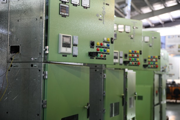 A row of industrial control panels painted in green with various dials, switches, and meters. The panels are aligned under a metal roof, and there are cables visible on one side. Brightly colored buttons in red, blue, yellow, and green are prominently placed on the panels.