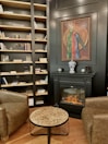 The interior of a cozy room features a well-stocked bookcase filled with books and decorative items against a dark wall. A sophisticated fireplace with a realistic flame sits below a framed painting. A small marble-top coffee table is placed between two brown leather armchairs, creating a comfortable sitting area. The wooden flooring adds warmth to the space.