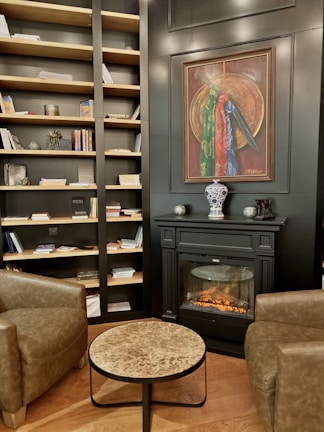 The interior of a cozy room features a well-stocked bookcase filled with books and decorative items against a dark wall. A sophisticated fireplace with a realistic flame sits below a framed painting. A small marble-top coffee table is placed between two brown leather armchairs, creating a comfortable sitting area. The wooden flooring adds warmth to the space.