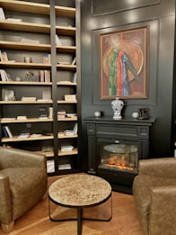 The interior of a cozy room features a well-stocked bookcase filled with books and decorative items against a dark wall. A sophisticated fireplace with a realistic flame sits below a framed painting. A small marble-top coffee table is placed between two brown leather armchairs, creating a comfortable sitting area. The wooden flooring adds warmth to the space.