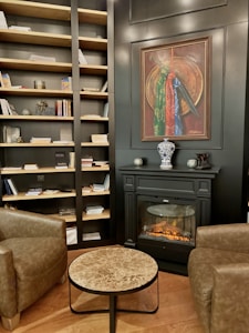 The interior of a cozy room features a well-stocked bookcase filled with books and decorative items against a dark wall. A sophisticated fireplace with a realistic flame sits below a framed painting. A small marble-top coffee table is placed between two brown leather armchairs, creating a comfortable sitting area. The wooden flooring adds warmth to the space.