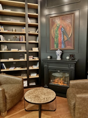 The interior of a cozy room features a well-stocked bookcase filled with books and decorative items against a dark wall. A sophisticated fireplace with a realistic flame sits below a framed painting. A small marble-top coffee table is placed between two brown leather armchairs, creating a comfortable sitting area. The wooden flooring adds warmth to the space.