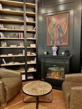 The interior of a cozy room features a well-stocked bookcase filled with books and decorative items against a dark wall. A sophisticated fireplace with a realistic flame sits below a framed painting. A small marble-top coffee table is placed between two brown leather armchairs, creating a comfortable sitting area. The wooden flooring adds warmth to the space.