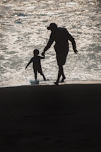 Silhouettes of an adult and a child walking hand in hand along the shore, with sunlight reflecting on the water.