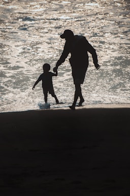 Silhouettes of an adult and a child walking hand in hand along the shore, with sunlight reflecting on the water.