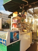 A small convenience store display with a variety of packaged snacks and goods organized on shelves. A freezer with Pepsi branding is in the foreground, illuminated by warm sunlight. The store's entrance is visible with glass doors and fluorescent lighting inside. Residential buildings and trees are seen in the background.
