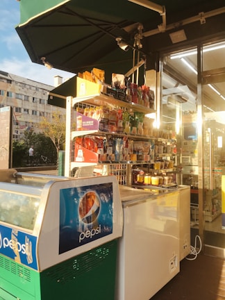 A small convenience store display with a variety of packaged snacks and goods organized on shelves. A freezer with Pepsi branding is in the foreground, illuminated by warm sunlight. The store's entrance is visible with glass doors and fluorescent lighting inside. Residential buildings and trees are seen in the background.