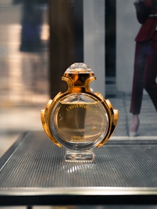 A perfume bottle with a luxurious gold and glass design is displayed on a sleek textured surface. The background is softly blurred, suggesting an indoor setting.