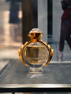 A perfume bottle with a luxurious gold and glass design is displayed on a sleek textured surface. The background is softly blurred, suggesting an indoor setting.