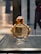 A perfume bottle with a luxurious gold and glass design is displayed on a sleek textured surface. The background is softly blurred, suggesting an indoor setting.