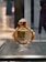 A perfume bottle with a luxurious gold and glass design is displayed on a sleek textured surface. The background is softly blurred, suggesting an indoor setting.