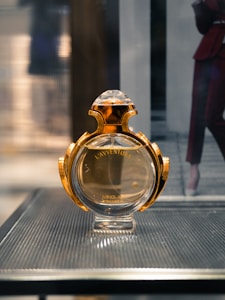 A perfume bottle with a luxurious gold and glass design is displayed on a sleek textured surface. The background is softly blurred, suggesting an indoor setting.