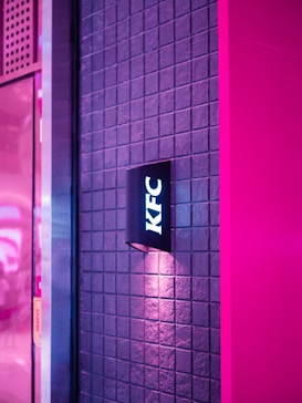 A wall with a grid pattern is illuminated by pinkish and purple lighting. A black sign with white letters spelling 'KFC' is mounted on the wall, casting a soft glow around it. The overall ambiance is modern and vibrant.