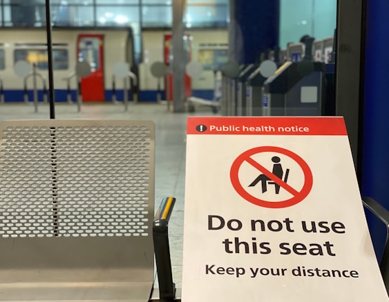 A public health notice indicating not to use a particular seat, with a red prohibition sign and the text 'Do not use this seat. Keep your distance'. The background shows part of a train station with a train and ticket barriers.