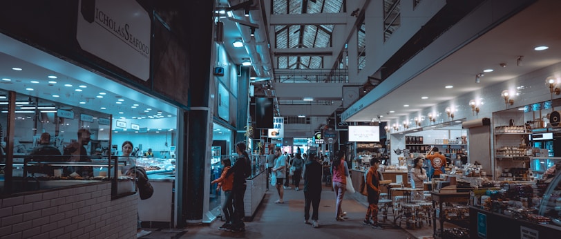 An indoor market scene with various stalls and shops lining both sides of a bustling hallway. People are seen shopping, browsing, and walking through the area. On the left, a seafood market is visible with a counter and employees inside. To the right, a deli or grocery section displays products under warm lighting. The ceiling features a lattice of windows, and the space is well-lit with overhead lights.
