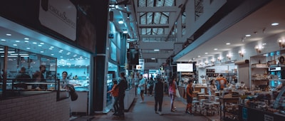 An indoor market scene with various stalls and shops lining both sides of a bustling hallway. People are seen shopping, browsing, and walking through the area. On the left, a seafood market is visible with a counter and employees inside. To the right, a deli or grocery section displays products under warm lighting. The ceiling features a lattice of windows, and the space is well-lit with overhead lights.