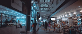 An indoor market scene with various stalls and shops lining both sides of a bustling hallway. People are seen shopping, browsing, and walking through the area. On the left, a seafood market is visible with a counter and employees inside. To the right, a deli or grocery section displays products under warm lighting. The ceiling features a lattice of windows, and the space is well-lit with overhead lights.