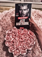 A person is holding a book titled 'Jack Hawthorne' by Ella Maise, which features a black and white portrait of a man on the cover. The background consists of a fluffy, pink textured surface. Beside the book, there is an arrangement of pink roses, creating a romantic and soft ambiance.