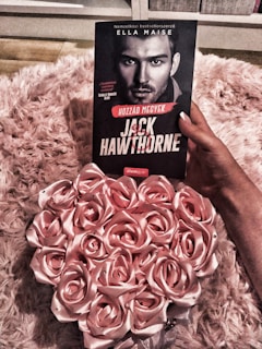 A person is holding a book titled 'Jack Hawthorne' by Ella Maise, which features a black and white portrait of a man on the cover. The background consists of a fluffy, pink textured surface. Beside the book, there is an arrangement of pink roses, creating a romantic and soft ambiance.