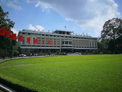 A large, white building with architectural design featuring vertical patterns stands prominently. Red flags are displayed along a pathway leading to the structure, which is set against a backdrop of a clear blue sky with scattered clouds. The foreground features a well-maintained green lawn, and there are people visible near the entrance.
