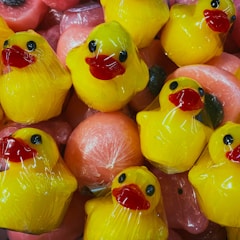 Bright yellow rubber ducks with red beaks are wrapped in clear plastic packaging. They are stacked closely together alongside round, pink objects that are also wrapped, giving the appearance of being in a display or for sale.