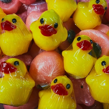Bright yellow rubber ducks with red beaks are wrapped in clear plastic packaging. They are stacked closely together alongside round, pink objects that are also wrapped, giving the appearance of being in a display or for sale.