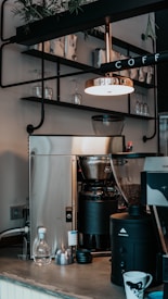 A cozy coffee shop setting with a modern coffee machine on a countertop, surrounded by glassware and coffee mugs. There is a metallic lamp hanging above, casting a warm light. Shelves stocked with coffee supplies and plants add to the ambiance.
