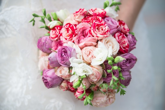 A vibrant bouquet of various flowers featuring shades of pink, red, white, and purple, accented by fresh green leaves. The flowers are tightly arranged, giving a lush and full appearance.